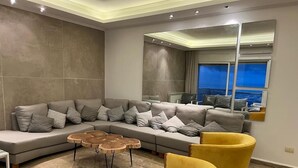 Ein modernes Wohnzimmer mit einem grauen Ecksofa, zwei gelben Sessel, einem runden Couchtisch und einem großen Spiegel, der den Raum reflektiert.