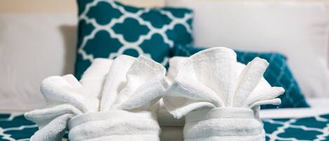 A bed with white and blue patterned bedding, two white towels shaped like bunny slippers, and a teal pillow with a white geometric pattern.