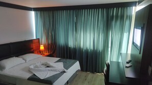 Quarto de hotel com cama, mesa de cabeceira com abajur, televisão e janela com cortinas.
