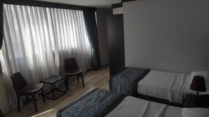 Quarto de hotel com duas camas, uma cadeira, uma mesinha e uma janela com cortinas leves.