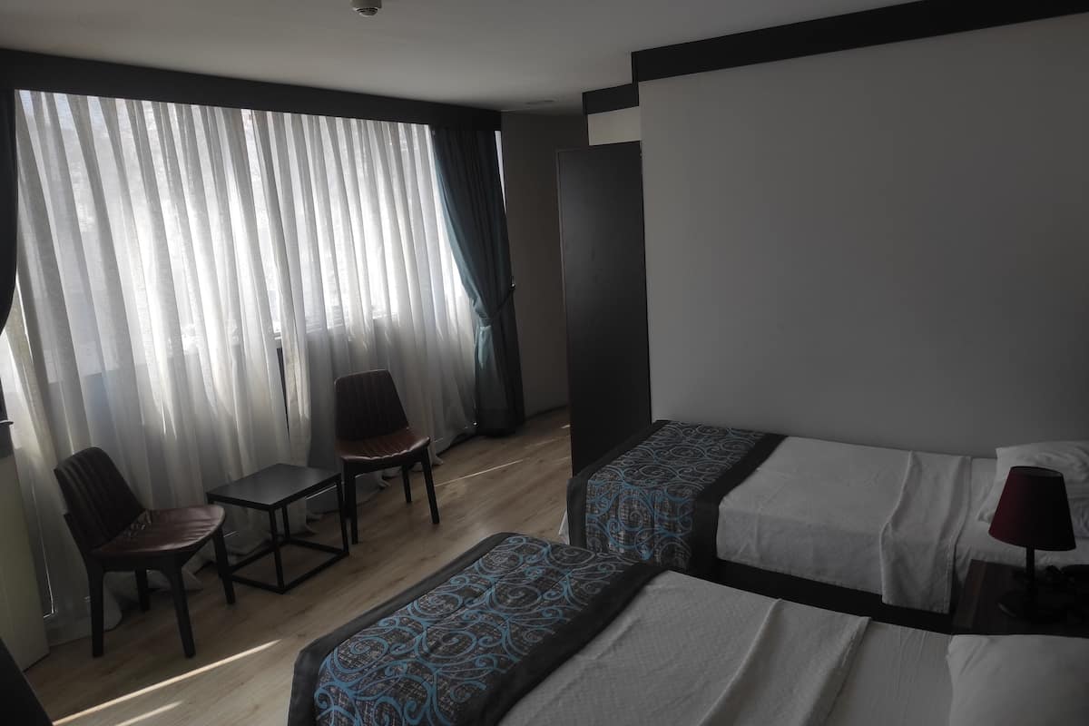 A hotel room with two beds, a chair, a small table, and a window with sheer curtains.