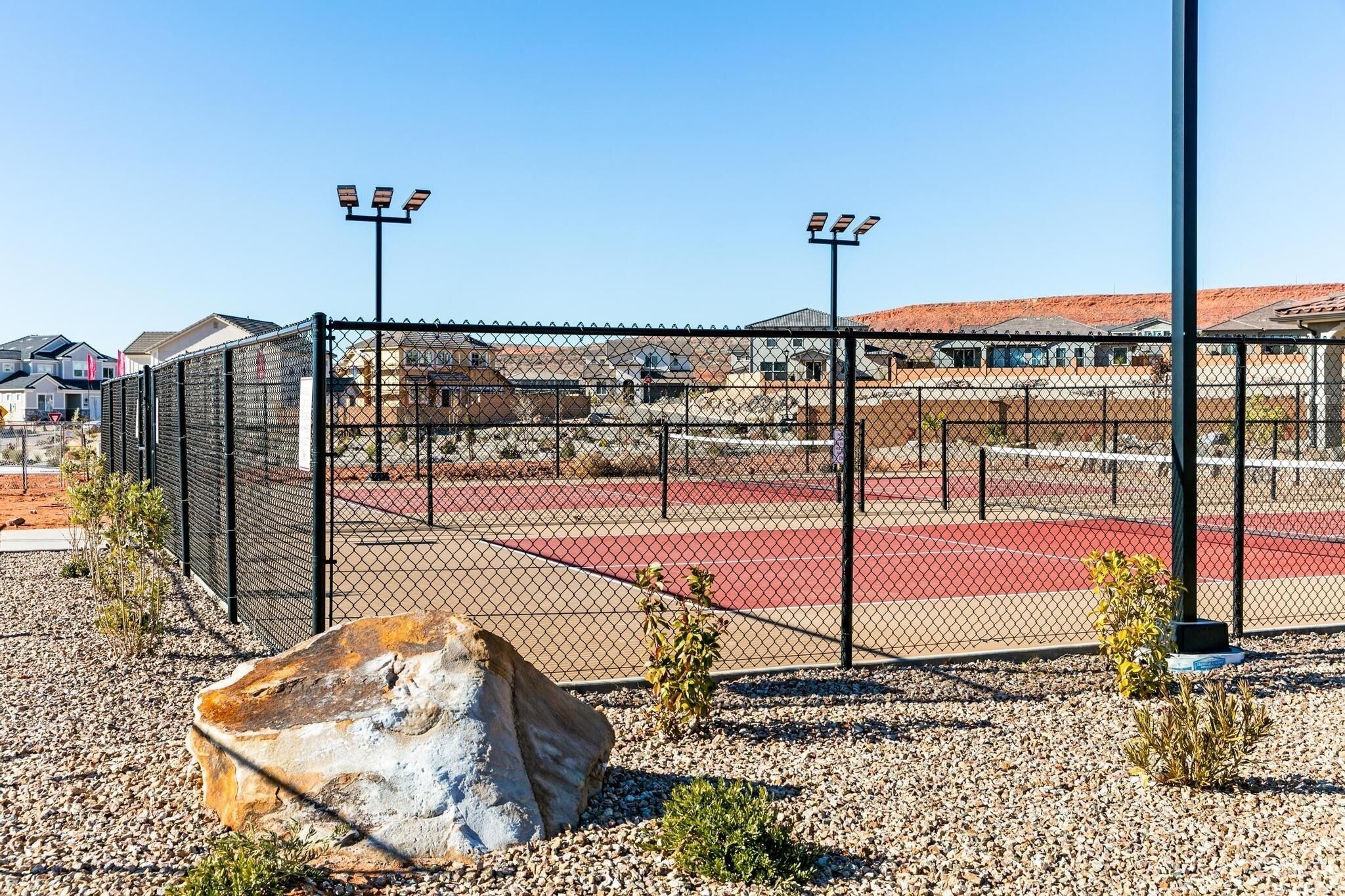 A fenced tennis court with a red surface, surrounded by a gravel area and small plants.