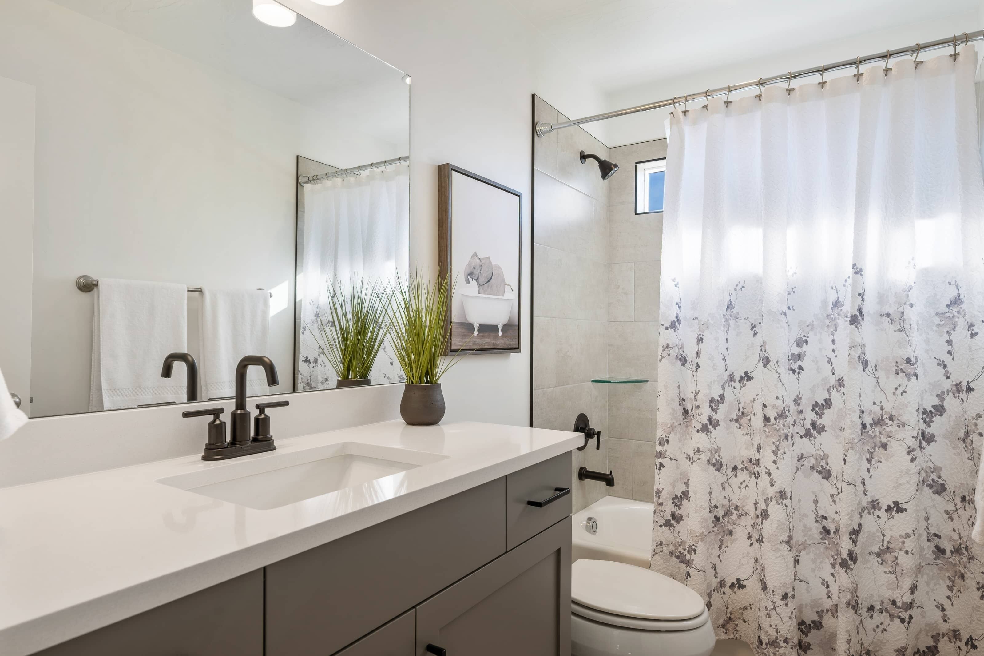 A modern bathroom with a white sink, two faucets, a mirror, a framed picture, a potted plant, a shower with a floral curtain, and a toilet.
