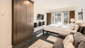 Une chambre moderne avec une grande armoire, un lit, un canapé, une télévision et un balcon donnant sur la forêt.