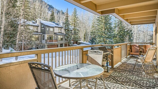 Un balcon avec une table et des chaises en verre, un barbecue et une vue sur des arbres et des bâtiments recouverts de neige.