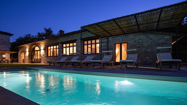 Zona junto a la piscina con sillones de descanso y un edificio de piedra con ventanas grandes.