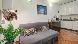 A living room with a sofa, two pillows, a potted plant, a painting, and a kitchen in the background. - Studio Saturno 23 in the City Center (Sauze d'Oulx)