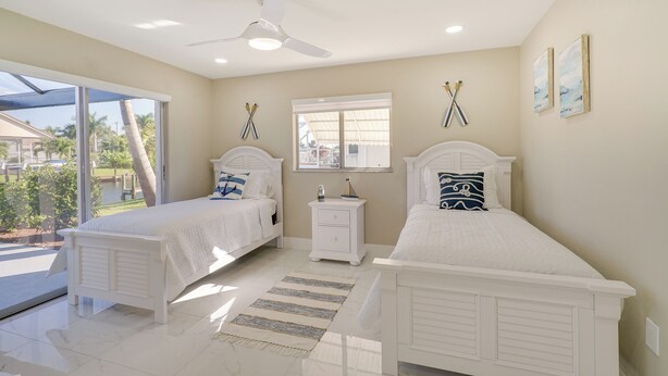A bedroom with two white beds, a ceiling fan, a nightstand, and a view of a pool and palm trees. - Screened Lanai w/ TV: Waterfront Cape Coral Home! (Cape Coral)