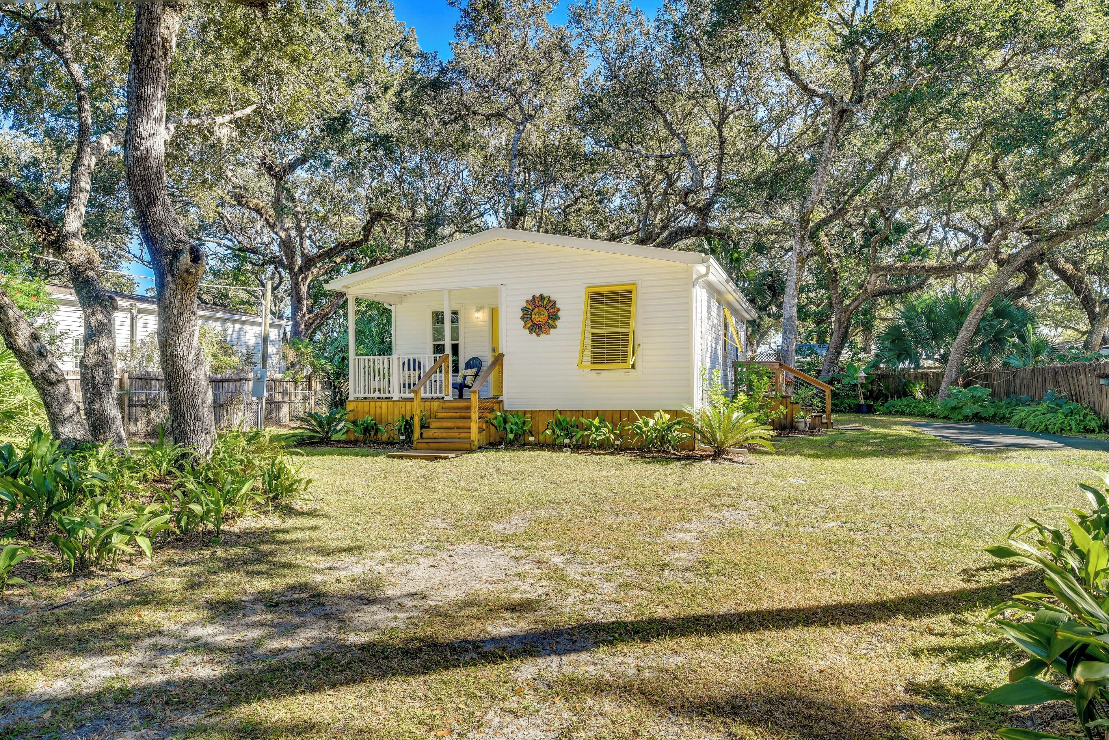 A small white mobile home with a porch and yellow shutters, surrounded by trees and a grassy yard.