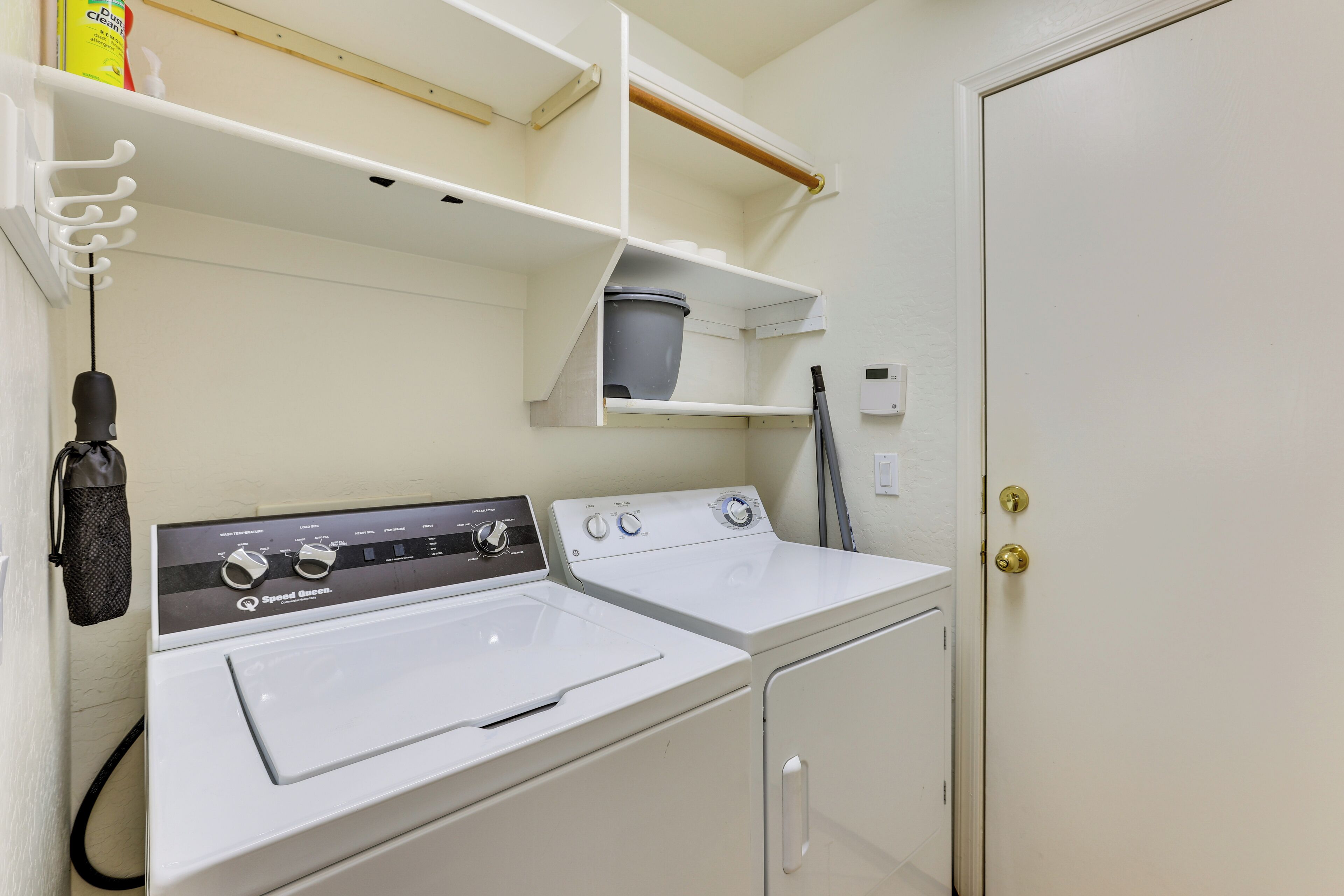A laundry room with two white front-loading washers and dryers, a shelf with a bucket, and a door with a brass handle.