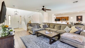 A spacious living room with a large sectional sofa, a coffee table, and a TV. A kitchen is visible in the background. - Spacious Surprise Home w/ Outdoor Pool + Game Room (Surprise)