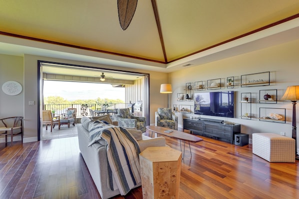 A spacious living room with a large TV, comfortable seating, and a balcony with a view.