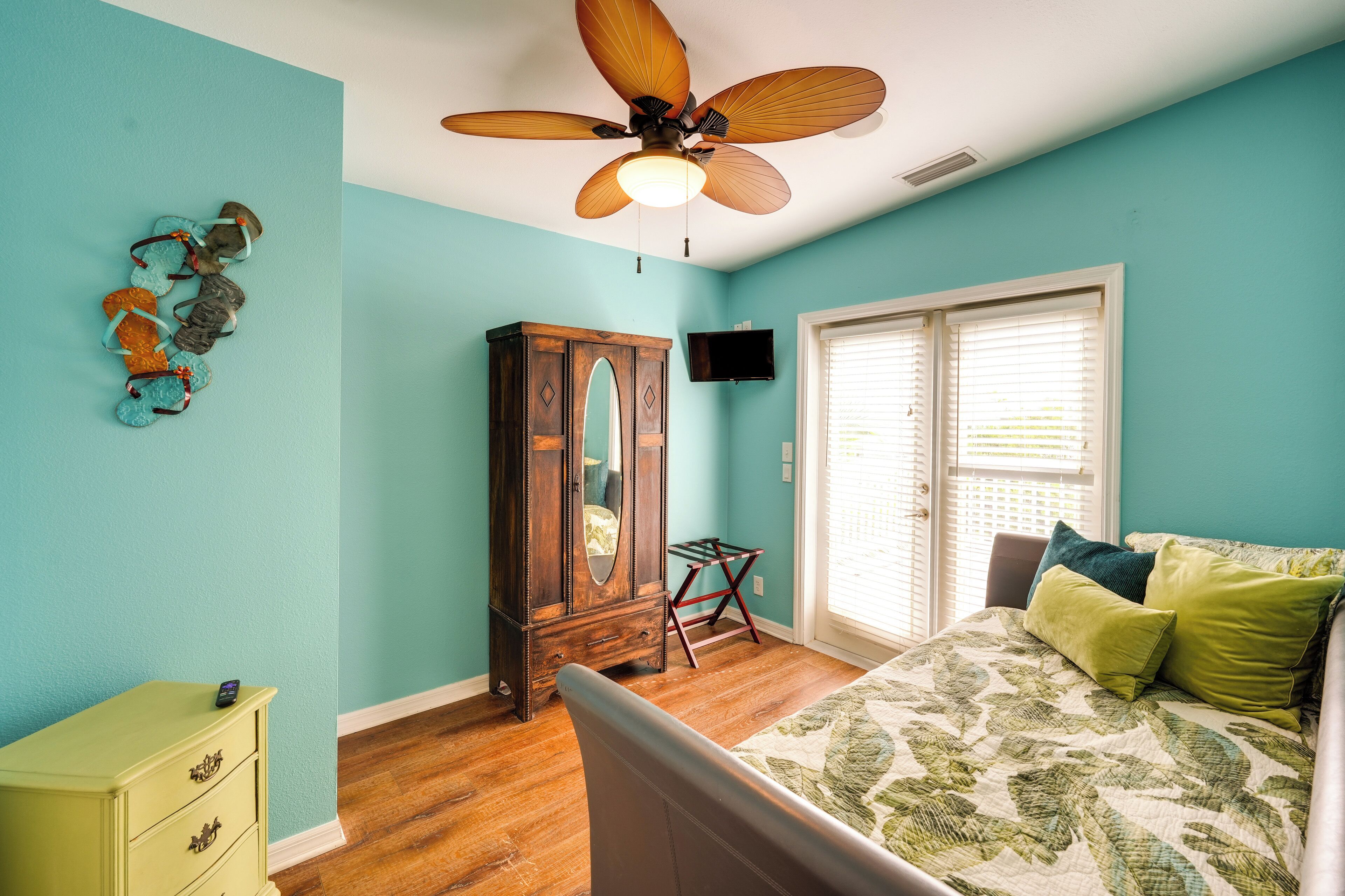 A bedroom with teal walls, a wooden dresser, a bed with a floral bedspread, a ceiling fan, and a window with blinds.