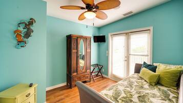 A bedroom with teal walls, a wooden dresser, a bed with a floral bedspread, a ceiling fan, and a window with blinds.
