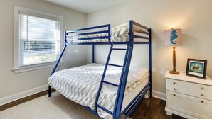 Un lit superposé avec une structure blanche et bleue, une commode blanche avec des tiroirs, une lampe décorée d’un motif représentant un hippocampe et une fenêtre avec des stores.