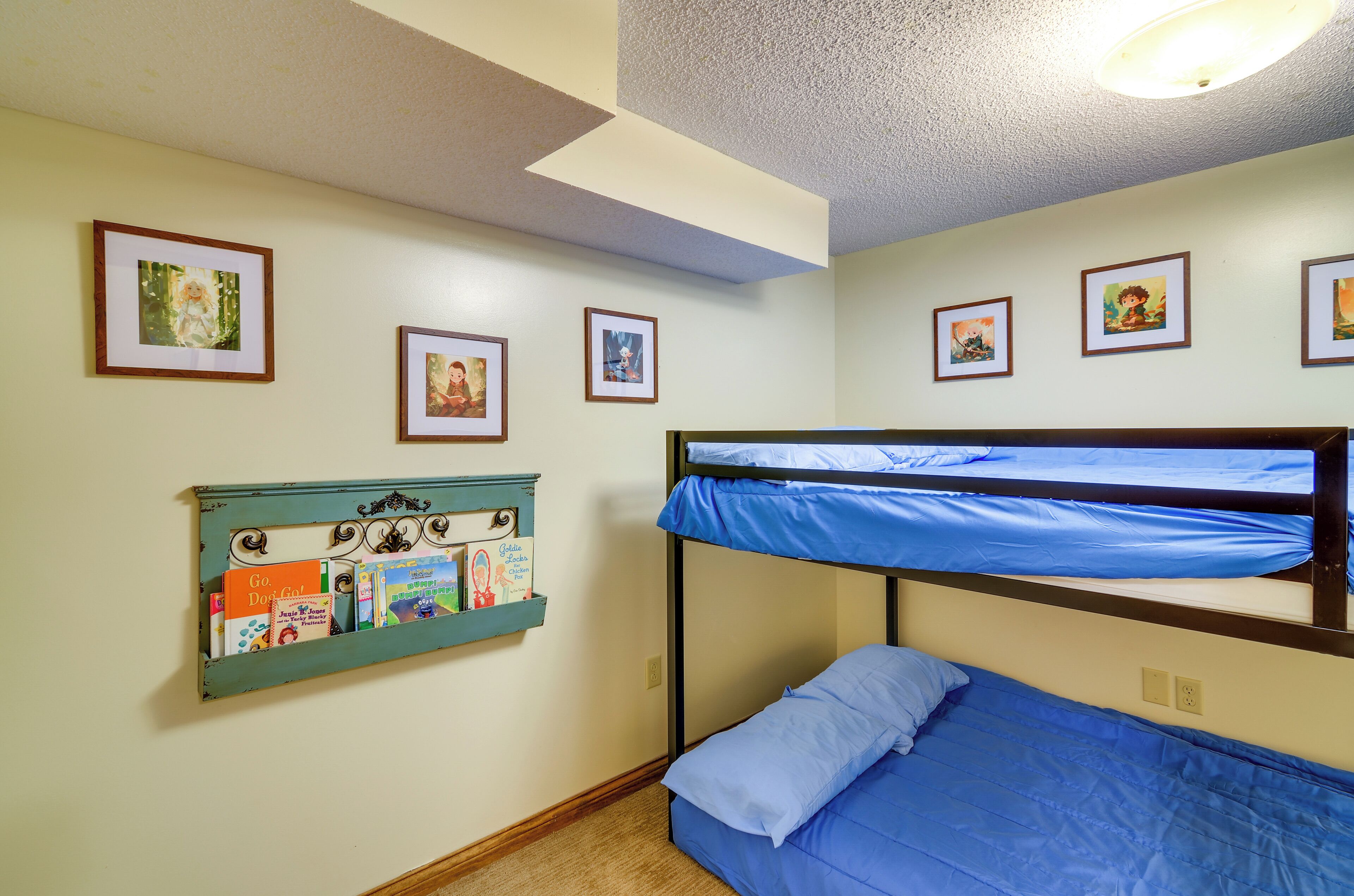 Une pièce avec des lits superposés, une étagère fixée au mur où sont rangés des livres pour enfants, et des tableaux encadrés accrochés au mur.