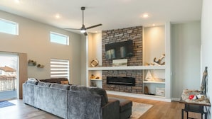 A modern living room with a gray sectional sofa, a stone fireplace, a flat-screen TV, and a ceiling fan. - Views of Lewis & Clark Lake - Modern Apt w/ Deck (Crofton)