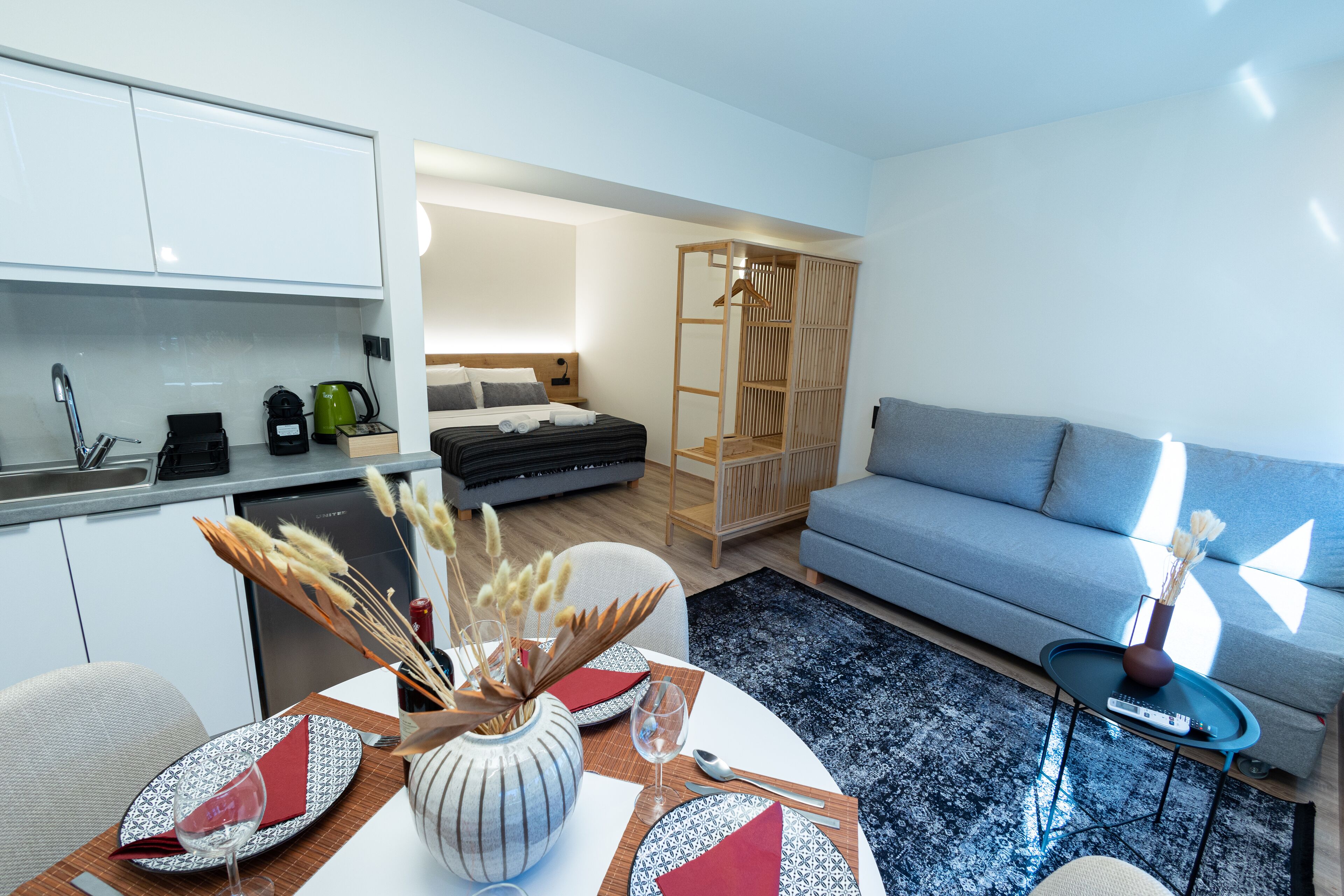 A modern living room with a sofa, a dining table set with plates and glasses, a coffee machine, and a kitchen area with cabinets and a sink.