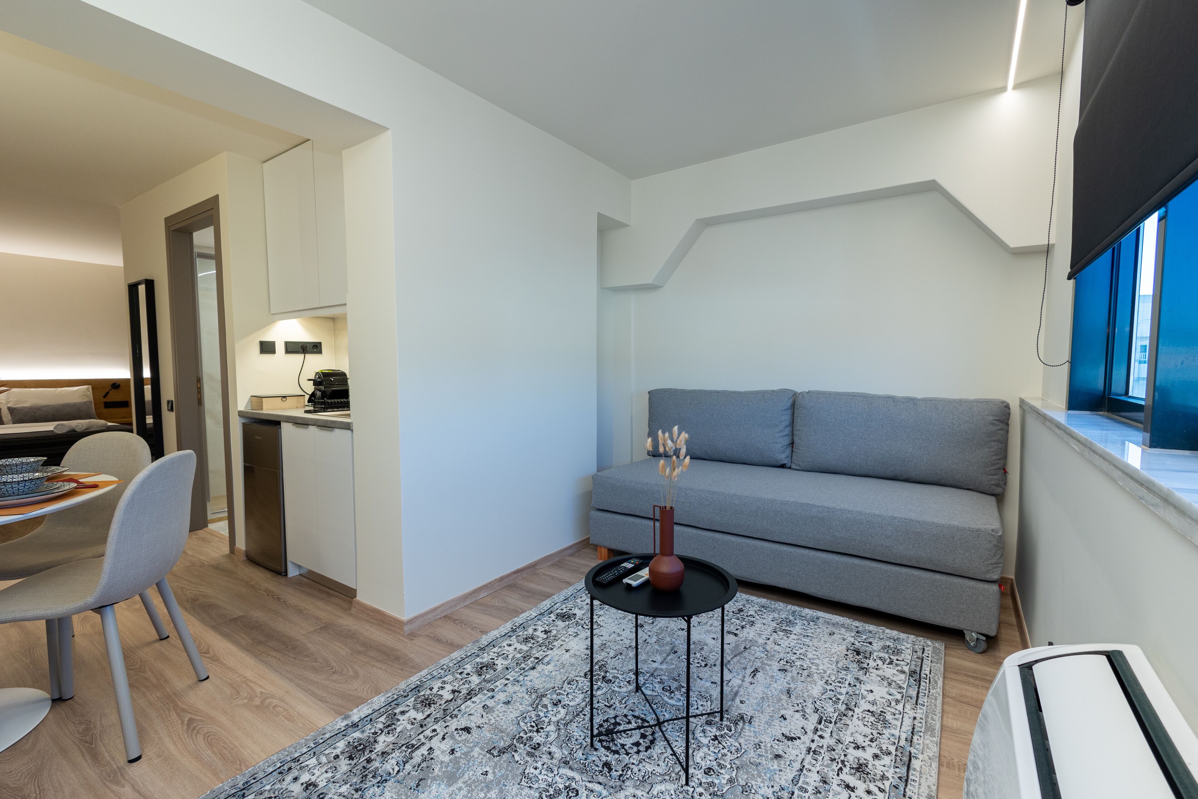 A modern living room with a grey sofa, a small round coffee table with a vase, a dining area with a table and chairs, and a kitchen area with a dishwasher.