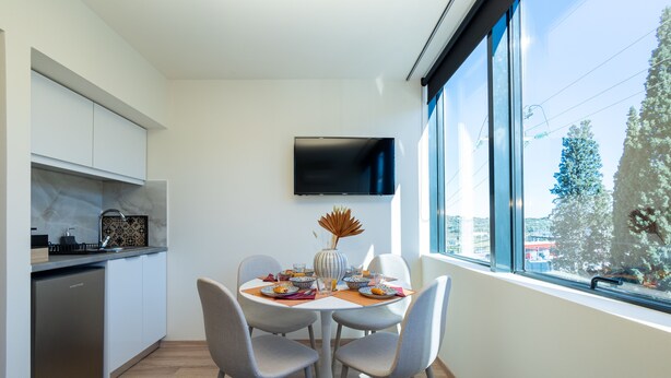A modern kitchen with white cabinets, a built-in dishwasher, and a dining area with a round table and four chairs. A flat-screen TV is mounted on the wall above the dining table. - Athens Airport Living spaces (Spata-Artemida)