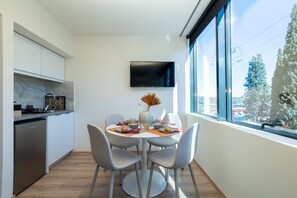 A modern kitchen with white cabinets, a built-in dishwasher, and a dining area with a round table and four chairs. A flat-screen TV is mounted on the wall above the dining table. - Athens Airport Living spaces (Spata-Artemida)