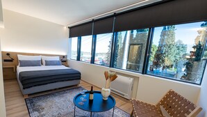 A modern bedroom with a bed, a small round table with a bottle and vase, a chair, and a large window with a view of trees and buildings. - Athens Airport Living spaces (Spata-Artemida)