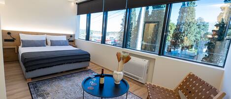 A modern bedroom with a bed, a small round table with a bottle and vase, a chair, and a large window with a view of trees and buildings.