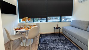 A modern living room with a grey sofa, a dining table with chairs, a black rug, and a large window with blinds. - Athens Airport Living spaces (Spata-Artemida)