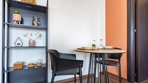 Una habitación moderna con piso de madera oscura, una mesa redonda con dos sillas, una estantería con objetos decorativos y una pared blanca con un detalle en color naranja.