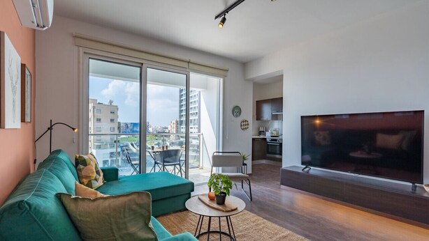 Una sala de estar moderna con un sofá color turquesa, un televisor de pantalla plana y un amplio ventanal corredero que da a un balcón.