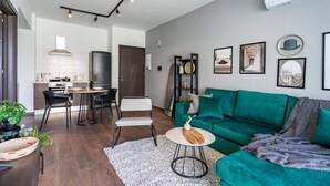 A modern living room with a teal sofa, a dining area with a round table, and a kitchen in the background. - Harbor 102: Stylish 1-Bedroom Apartment in Larnaca (Cyprus - Larnaca)