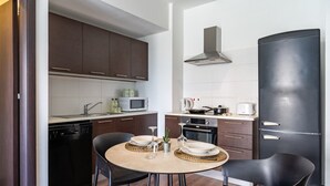 A compact kitchen with dark cabinets, a microwave, a stove, and a refrigerator. There is a dining table with chairs and a small potted plant. - Harbor 102: Stylish 1-Bedroom Apartment in Larnaca (Cyprus - Larnaca)
