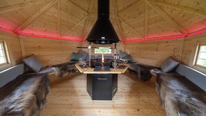 Interior acogedor de una cabaña con chimenea central, paredes de madera y techo alto.