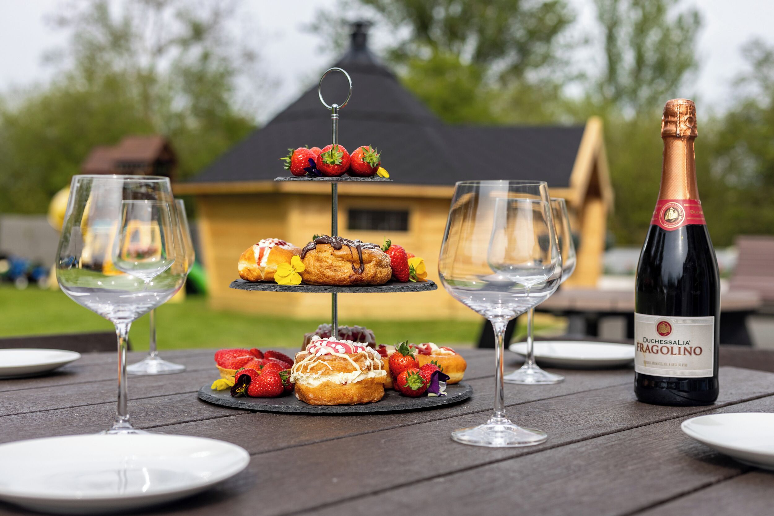 A picnic setup with a three-tiered dessert stand, two wine glasses, and a bottle of Duchessalia Fragolino wine on a wooden table.