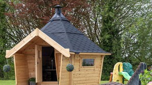 Una casita de juegos de madera con techo a dos aguas y chimenea, ubicada en un césped con árboles al fondo.