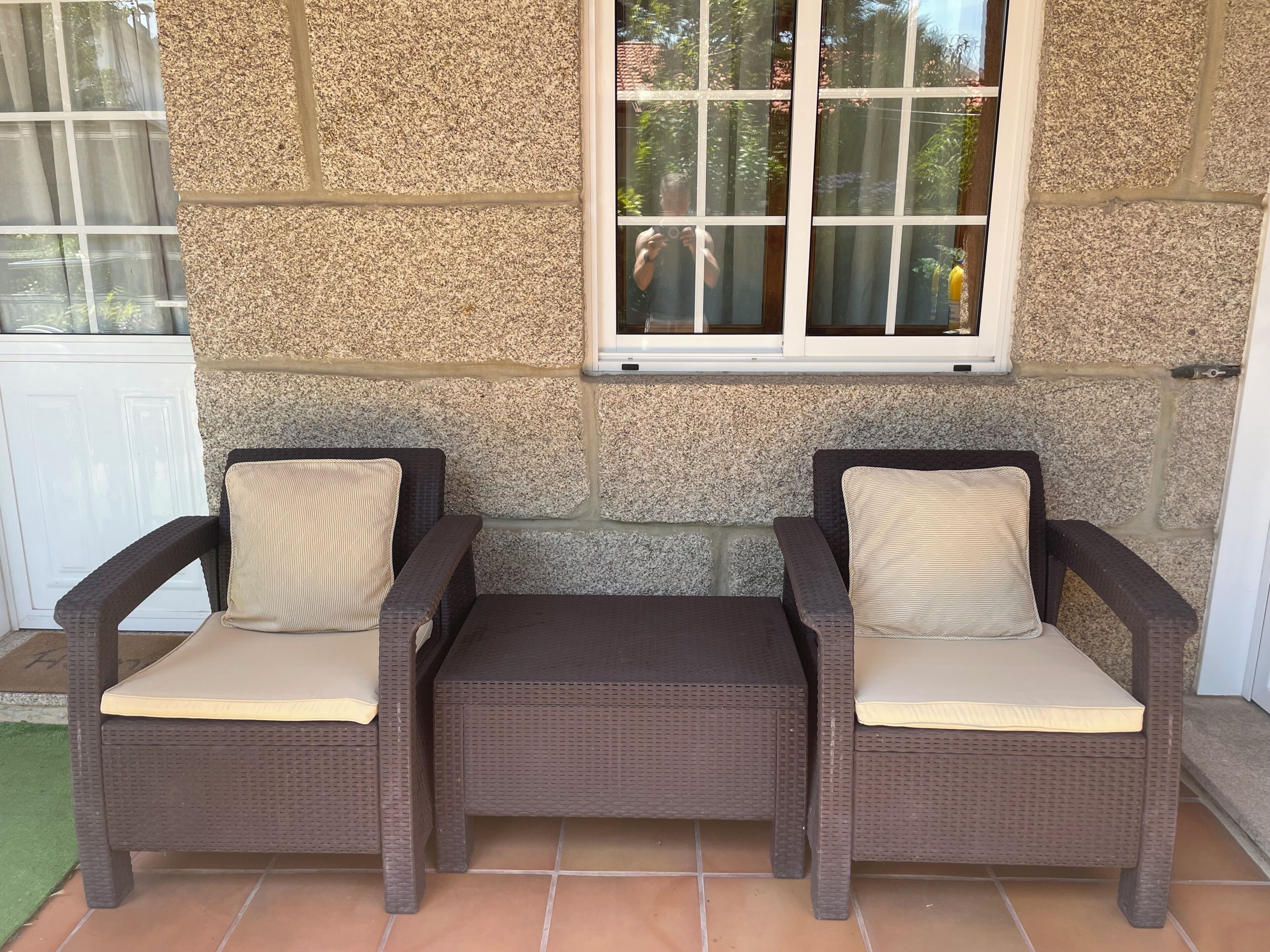A patio area with two wicker chairs and a bench, a stone wall, and a window with a view of greenery.