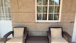 A patio area with two wicker chairs and a bench, a stone wall, and a window with a view of greenery.