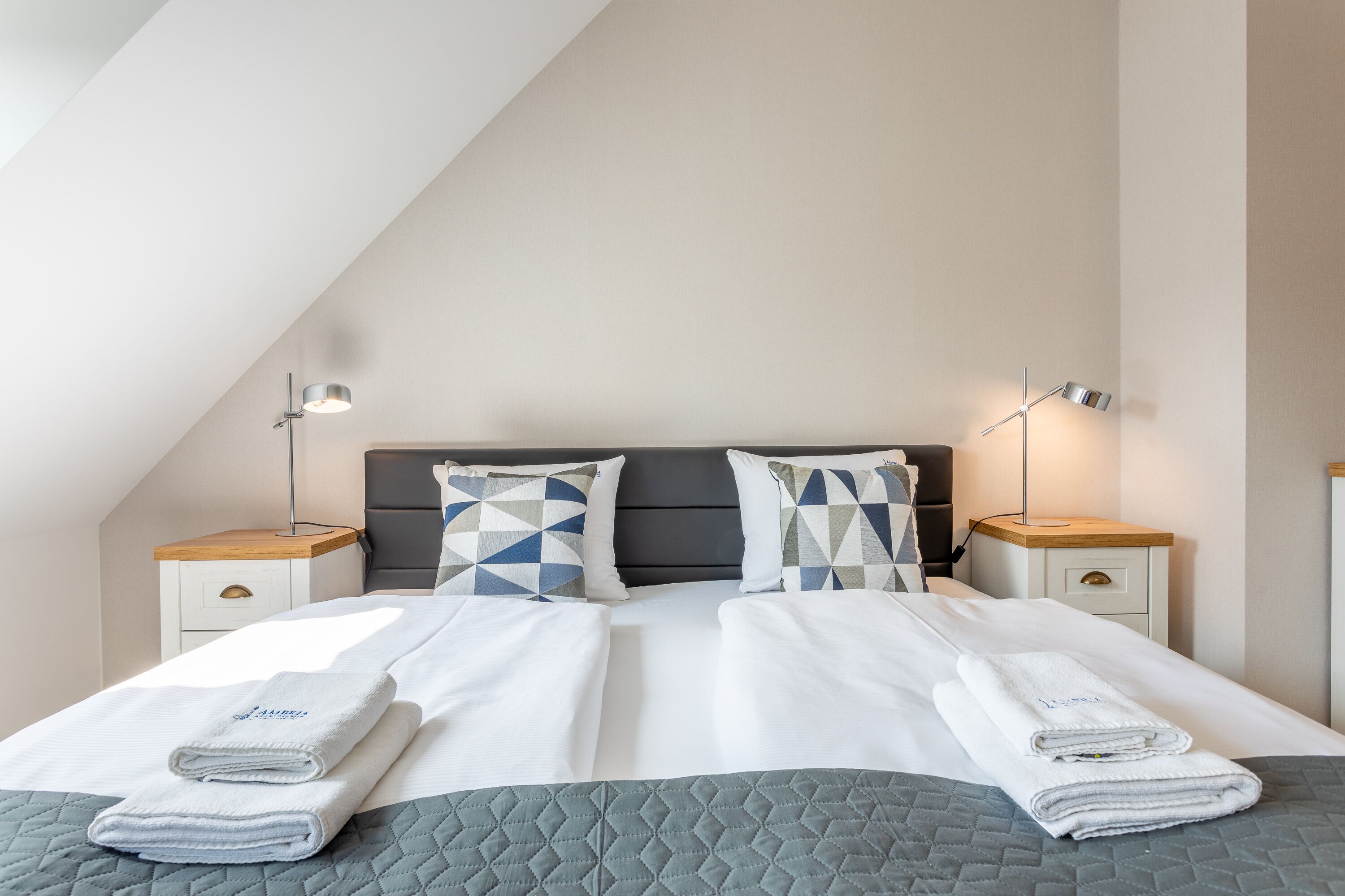 A neatly made bed with folded towels, a geometric patterned pillow, and bedside lamps on a white nightstand.