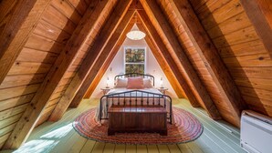 An attic bedroom with a wooden sloped ceiling, a bed with a headboard, a bedside table, a lamp, and a window with a view of trees. - A-Frame of Mind - Time to Kick Back and Relax (Demorest)