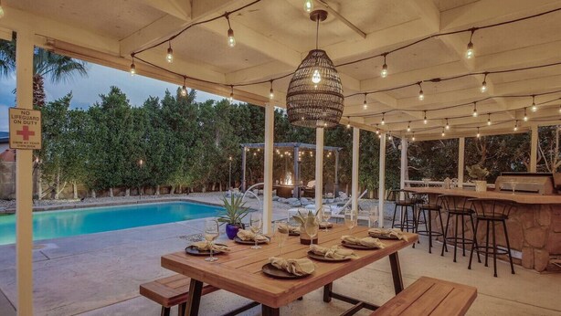 Une terrasse extérieure avec une piscine, des tables en bois disposées pour les repas, des guirlandes lumineuses et un coin bar.
