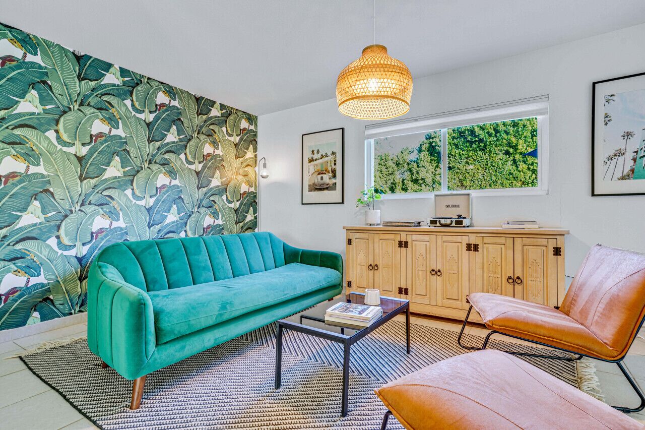 Een woonkamer met een smaragdgroene bank, een bruine leren fauteuil, een houten kast en een groot raam met uitzicht op groen.