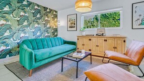 Un salon avec un canapé de couleur turquoise, un fauteuil en cuir marron, un meuble en bois et une grande fenêtre donnant sur un paysage verdoyant.
