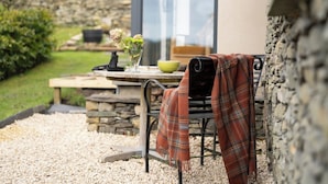 Un mur de pierres, une table avec un bol, et une chaise recouverte d’une couverture à carreaux.