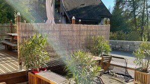 Ein Außenbereich mit einer Terrasse, Holzmöbeln, Topfpflanzen und Blick auf ein Haus im Hintergrund.