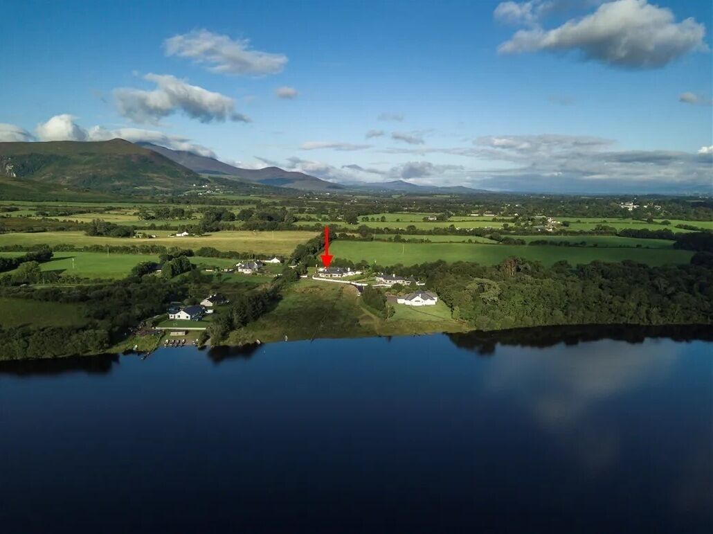 An aerial view of a lakeside property with multiple buildings, a red arrow indicating a point of interest, and surrounding green fields and hills.