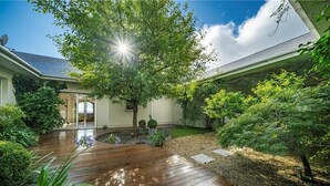 A modern house with a wooden deck, a covered patio, and a garden with various plants and a tree.