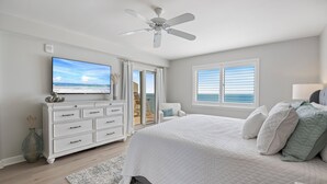 Ein Schlafzimmer mit einem großen Bett, einem Deckenventilator, einem Fernseher und Blick auf den Strand durch das Fenster.