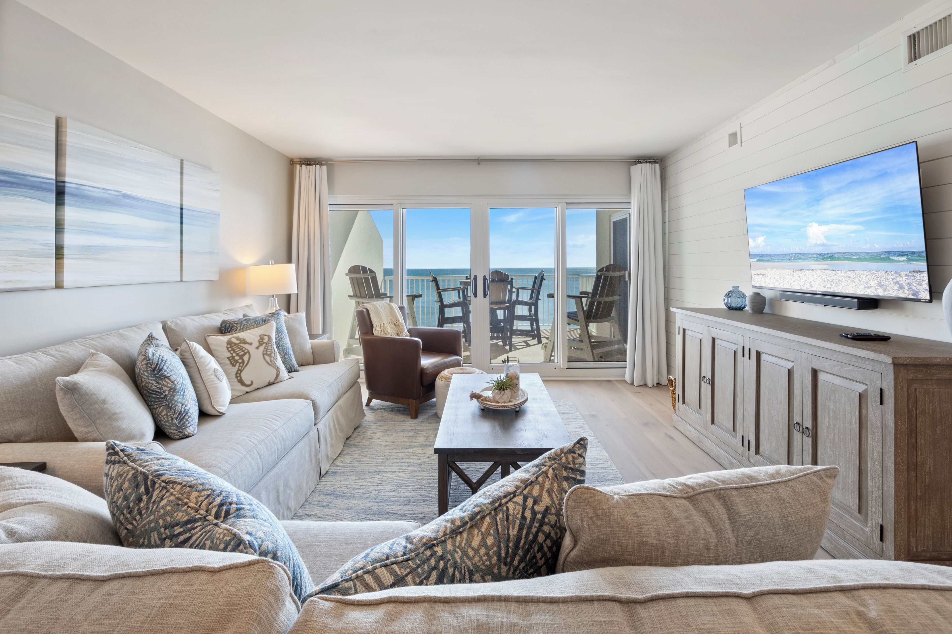 A spacious living room with a large television, a sofa set, and a view of the ocean through the glass doors.