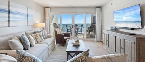 A spacious living room with a large television, a sofa set, and a view of the ocean through the glass doors.