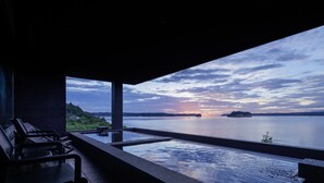 Un área de piscina con vistas al atardecer sobre el agua, sillones de descanso y una estructura de piedra.